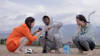 Three people crouching outdoors around a camera tripod, one holding a phone.