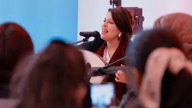 A woman singing into a microphone while playing a stringed instrument, with audience members visible in the foreground.