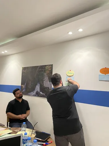 A man points to a sticky note reading "Motivation" on a wall while another man sits at a table watching, in a room with a photo of a waterfall.