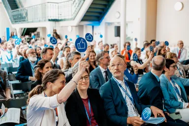 A seated audience in a hall holds up blue thumbs-up signs during a session, with a staircase visible in the background.