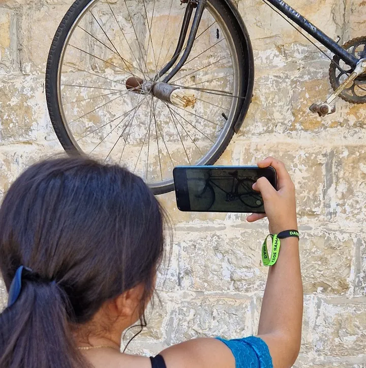 A person photographs an old bicycle wheel mounted on a stone wall using a smartphone.