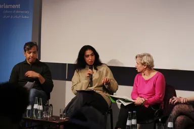 Three people sit on a panel stage, a woman in the middle speaking into a microphone, a banner partly showing "arlament cultures of democracy" text, water bottles on a table in front.
