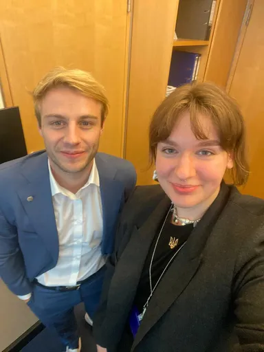 Two people pose for a selfie in an office, with wooden cabinet doors and binders on shelves behind them.