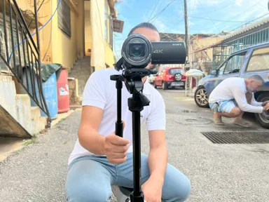 A man crouches outdoors filming with a video camera on a monopod while another man crouches beside a car in the background.