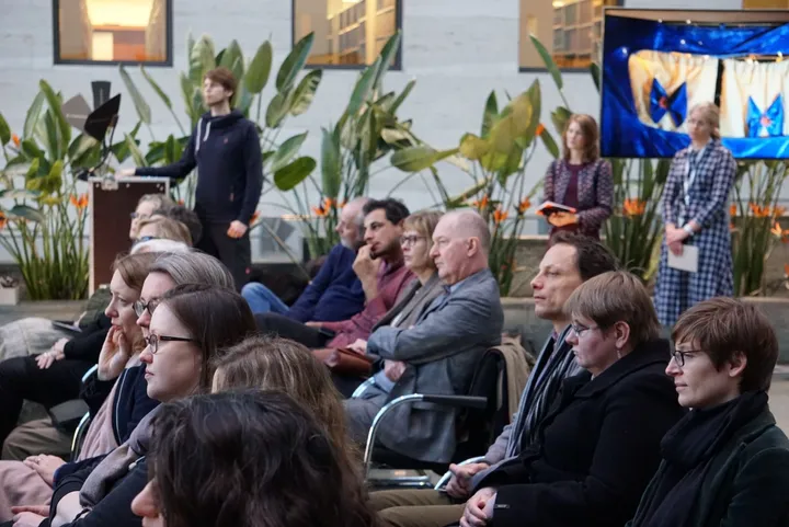 A group of seated people listening to a man standing beside a lectern, with a blue banner on a screen and potted plants in the background.