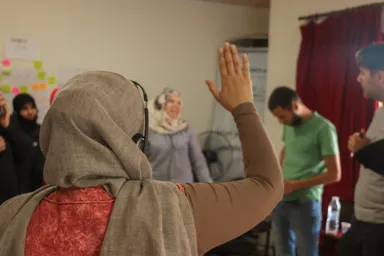 A person raises a hand in a room with a fan and papers on the wall, while two men and a woman stand nearby beside a red curtain.