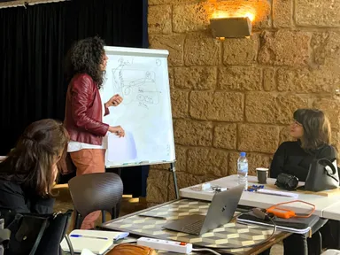 A woman in a red jacket sketches on a flip chart while two women sit and watch in a stone-walled room with a laptop on the table.