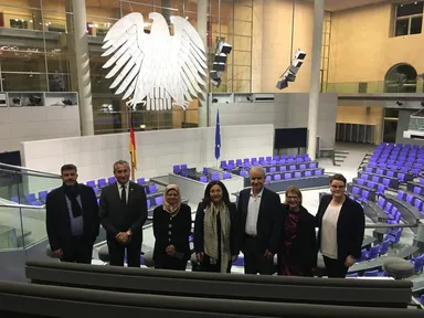 Seven people pose for a group photo in a parliamentary chamber beneath a large eagle emblem, with rows of empty blue seats and flags behind them.