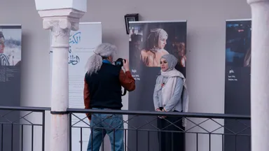 A man photographing a woman posing in front of exhibition banners showing portrait photographs with biographical captions, behind a railing.