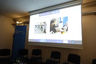 A projection screen displays a slide titled "What art can do" with two photographs and the text "The projects are funded by the German Federal Foreign Office", next to a banner reading "elbarlament" and rows of empty chairs.