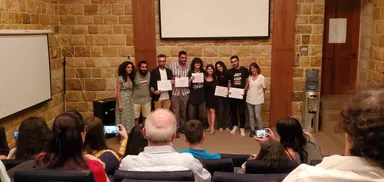 A group of people stand on a stage holding certificates while an audience films and photographs them.