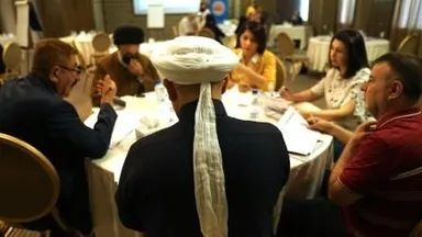 A person wearing a white turban sits with their back to the camera at a round table where several people talk and take notes.
