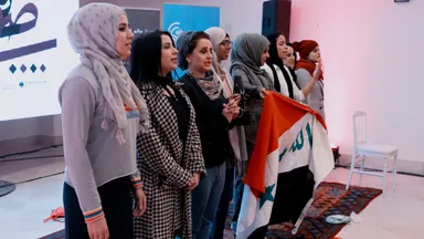 A row of women stand together, one holding a folded flag with red, white and black stripes and green Arabic script, in front of a stage backdrop with Arabic calligraphy.