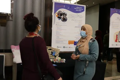 Two women wearing face masks talk beside a poster reading "We Are There for Each Other" and "نحن لبعض", with a woven bag and handicrafts on a table.