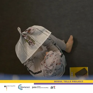 An overhead view of a person sitting on a floor holding an open newspaper with Arabic-script text over their face, above a "MOSUL TELLS PROJECT" banner.