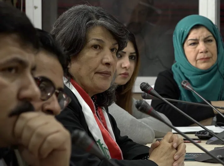 A group of people sits at a table with microphones, one woman wearing a teal headscarf.