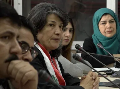 A group of people sits at a table with microphones, one woman wearing a teal headscarf.