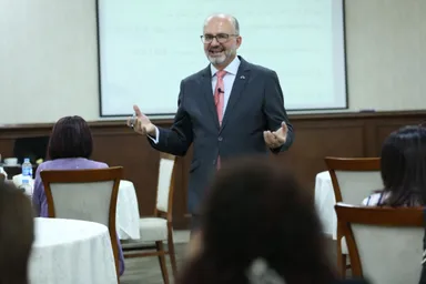 A man in a suit and pink tie gestures while speaking to a seated audience, with a screen behind showing partial text including "BAINA" and "WATNA".