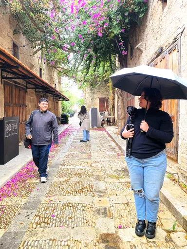 A woman holds an umbrella and carries camera equipment on a tripod in a flower-draped alley, with two people walking behind her.