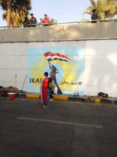 A person stands before a wall mural showing a silhouetted figure holding a flag inside a radiating sun design, with the word "IRAQ" and Arabic text painted beside it, while others watch from a bridge above.