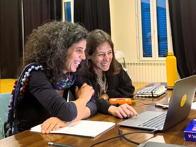 Two women laugh together while looking at a laptop screen at a table.