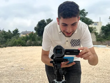 A man smiling while adjusting a Sony 4K camcorder mounted on a tripod outdoors near trees and buildings.