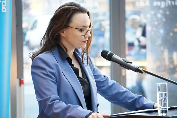 A woman speaking into a microphone at a lectern, wearing glasses and a blue blazer.