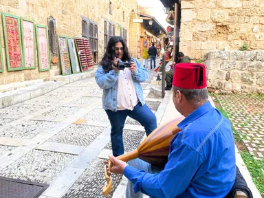 A woman films with a camcorder in a stone alleyway while a man in a red fez plays a stringed instrument in the foreground.