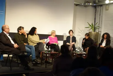 Seven people sit on a panel stage in front of a blank projection screen, a potted plant, and a white brick wall, one woman speaking into a microphone, water bottles on tables, an audience in the foreground.
