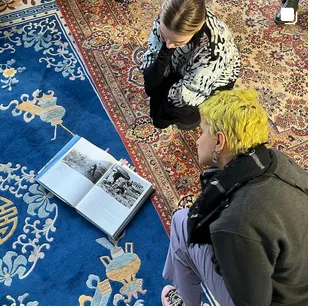 Overhead view of two people looking at an open book showing black-and-white photographs, placed on a blue and red patterned rug.