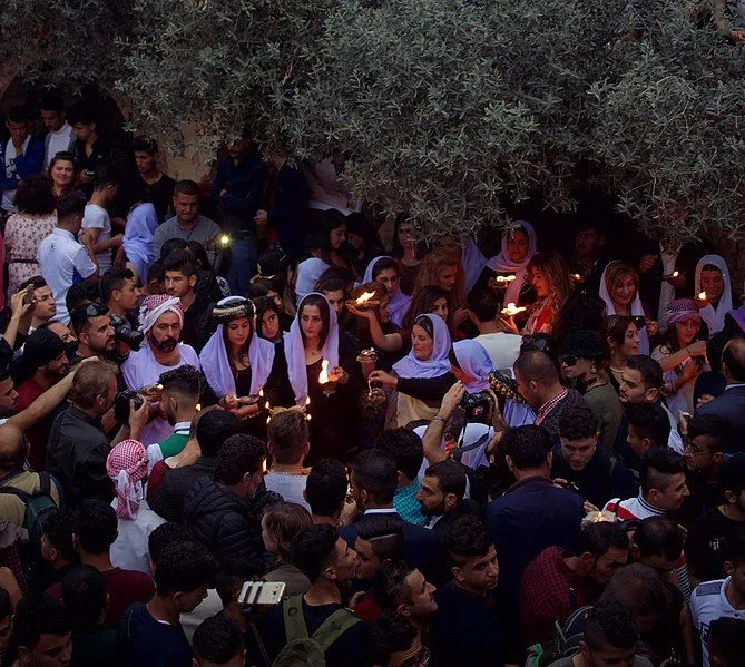 A crowd gathers outdoors at dusk under an olive tree, with several people in traditional dress holding lit candles.