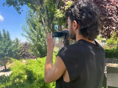 A man films a sunny garden with a handheld camera, seen from behind, with trees and a building in the background.