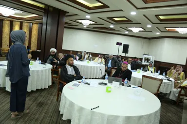 A woman wearing a headscarf stands and speaks at a conference with several round tables of seated attendees, including men in turbans and keffiyehs.