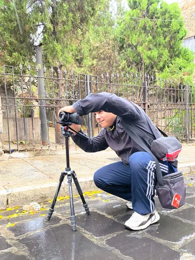 A man crouches operating a camcorder on a tripod on a wet street beside an iron fence.