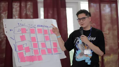 A man speaking into a microphone while pointing at a flip chart covered with a grid of pink sticky notes and handwritten notes reading "Что?".