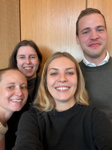 A group of four people smiling together for a selfie in front of a wood-paneled wall.