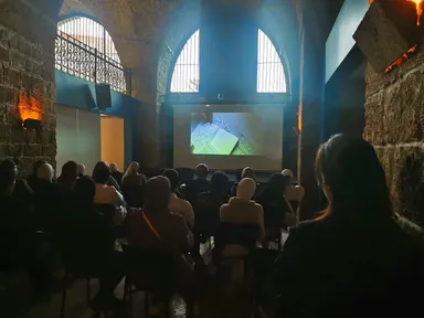 An audience sits in a dim vaulted hall watching a video projected on a screen that shows an open notebook.
