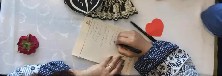 Overhead view of a hand writing Arabic text on a card next to a red paper heart, a rose, and a fan printed with a broken-heart symbol.