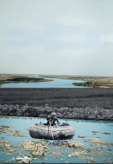 A person paddles a round boat across water littered with floating waste, beside a dark barren landscape and a river in the distance.