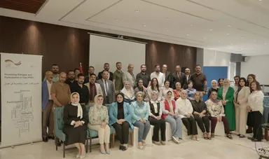 A large group of people pose in a conference hall beside a banner reading "Promoting Dialogue and Participation in Iraq (PDP)" and a word cloud including "Democracy" and "Dialogue".