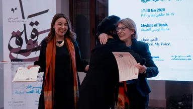Two women embrace on stage while holding certificates, in front of a screen reading "17-18 Jan 2020" and "Medina of Tunis" and a banner reading "My Pen. My Voice.".