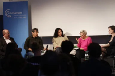 Five people sit on a panel stage in front of a blank projection screen and a banner reading "elbarlament cultures of democracy" with Arabic text, an audience seated in the foreground.