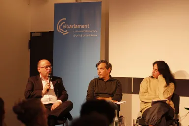 Three people sit on a panel stage in front of a blank projection screen and a banner reading "elbarlament cultures of democracy" with Arabic text, a man speaking into a microphone.