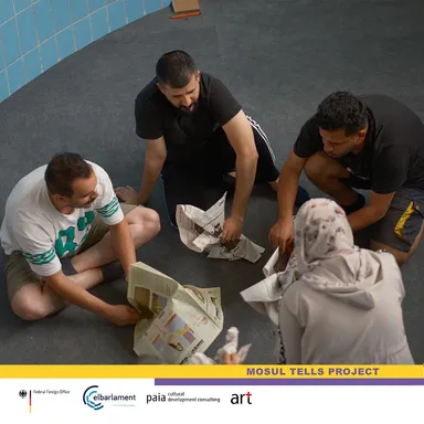 Four people crouching on a floor unfolding newspapers, above a banner reading "MOSUL TELLS PROJECT".