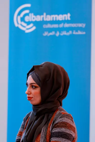 A woman wearing a dark headscarf and a striped cardigan stands in profile in front of a blue banner reading "elbarlament / cultures of democracy" with Arabic text below.