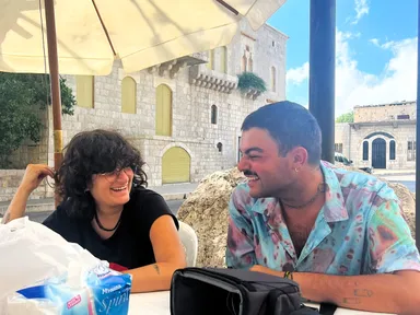 A woman and a man laugh together at an outdoor café table with a stone building in the background.