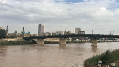 A river with a bridge, mosque minarets and domes, and a city skyline under a cloudy sky.