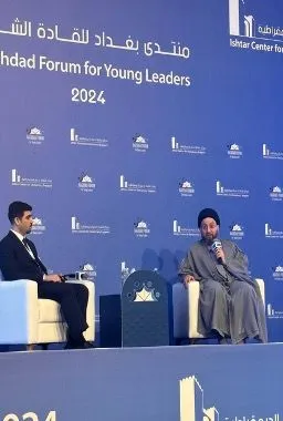 Two men sit on a stage in front of a banner reading "Baghdad Forum for Young Leaders 2024", one speaking into a microphone.