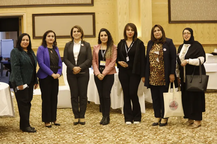 A group of seven women standing side by side in a conference room, some holding bags.