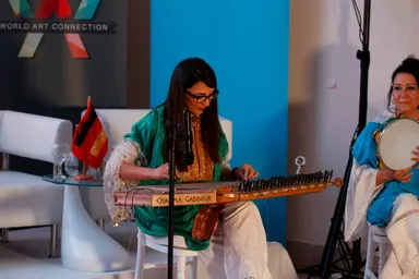 A woman in a green and gold embroidered outfit plays a stringed instrument with a nameplate, beside a woman playing a tambourine, in front of a banner reading "WORLD ART CONNECTION" and a flag on a table.